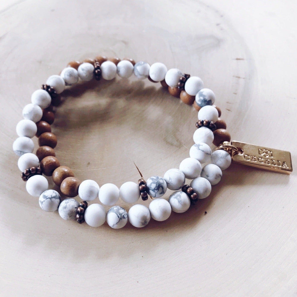 A stretch syle double-wrap bracelet made of white gray howlite, and brown wood, beads with copper accent beads, and a gold-tone logo tag, displayed on a light tan background.