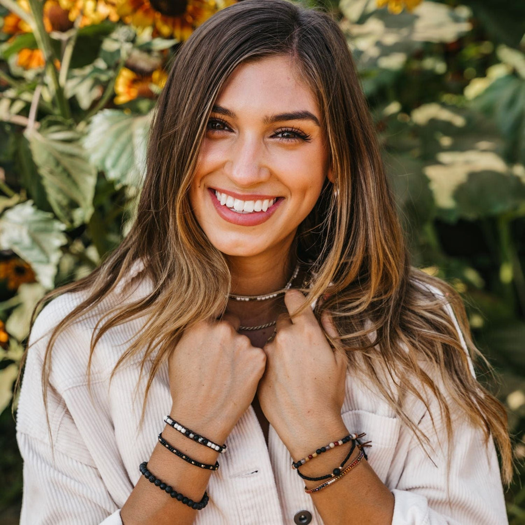 Portrait of a young woman with long brown hair, brown eyes and big smile, in a white corduroy jacket, wearing three bracelets on each wrist in shades of black, white, red, and green, with blurred sunflower plants in background.