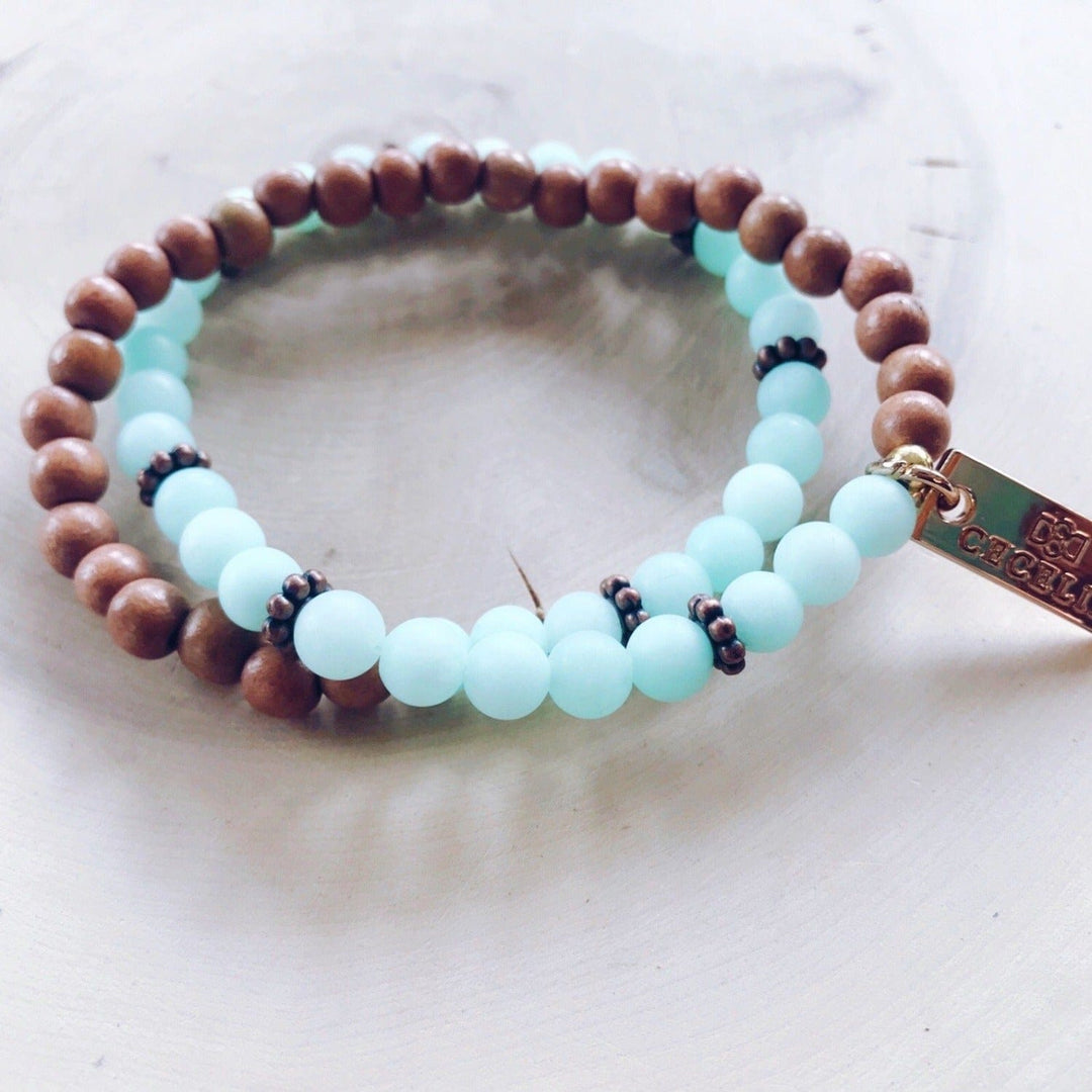 A stretch syle double-wrap bracelet made with light blue jade, and brown wood, beads with copper accent beads, and a gold-tone logo tag, displayed on a light tan wood background.