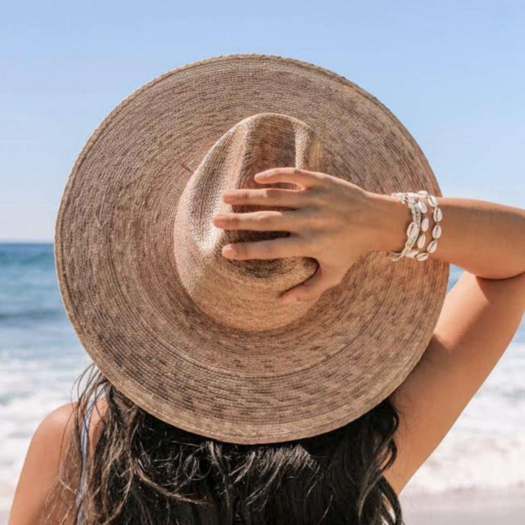 Close-up of the back of a woman's head with the blue sky and blurry ocean in the background, with long dark brown hair wearing a straw sun hat, with her hand holding the back of the hat and multiple cowry sea shell bracelets on her wrist, including the Kai natural white colored raided cowry bracelet.