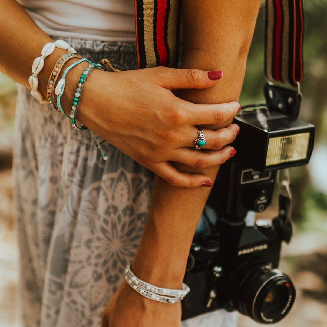 Close-up of the side of a woman's torso with her hand laying across her opposite arm, dresses in gray and white print flowy pants and white top, with a camera and carry strap over her shoulder, wearing multiple beaded bracelets on each wrist, including the braided blue cowry shell bracelet.