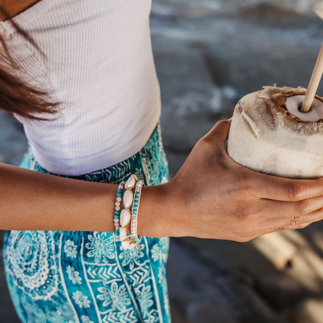Image of a young woman's torso and arm holding a coconut with a straw, wearing a white tank top with turquoise blue print pants, and three bracelets on her wrist, one with white sea shells, a bead bracelet and a bead wrap bracelet, both in shades of white and turquoise.