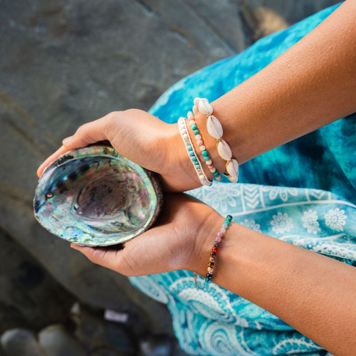 close-up of a young woman's hands holding an abolone shell wearing turquoise blue print flowy pants, and three bracelets on one wrist, one is a sea shell bracelet, one is a bead bracelet, and one is a wrap, both in pastel shades of blue, pink, and white, and a multicolored bead bracelet on the other wrist, with a gray rock background.