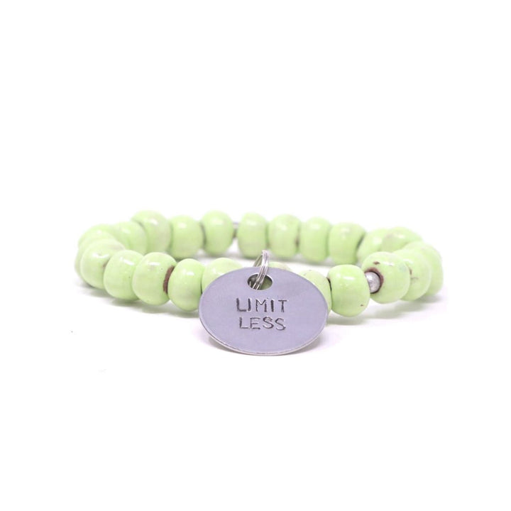 Close-up of a stretch style bracelet made with pale green clay beads and silver accent beads, with a silver metal charm with the inscription 'LIMITLESS', displayed on a white background.