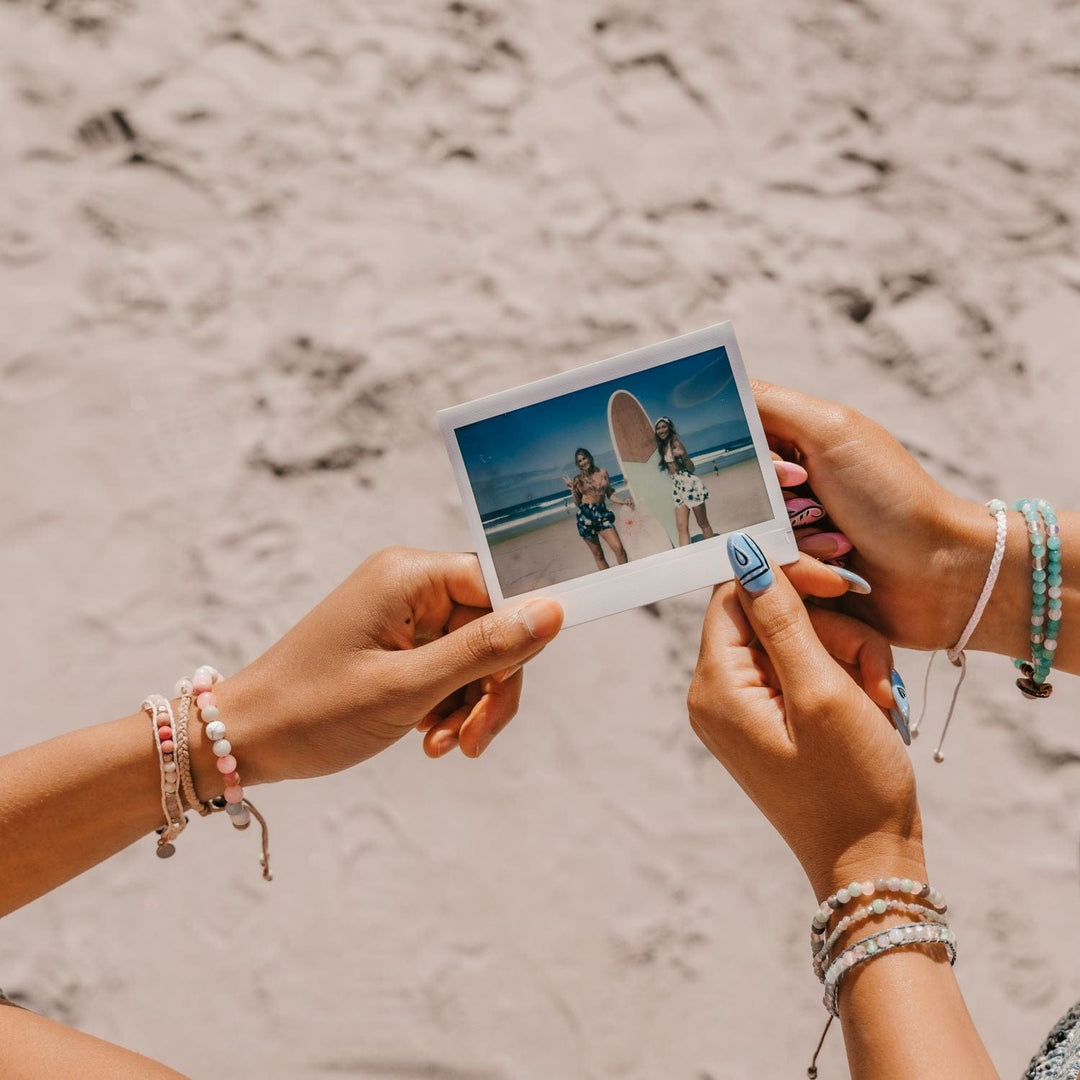 Close-up of two women's hands wearing multiple bracelets on each wrist, with blurred beach sand in the background, holding a polaroid photo of two women at the beach standing next to a surf board.