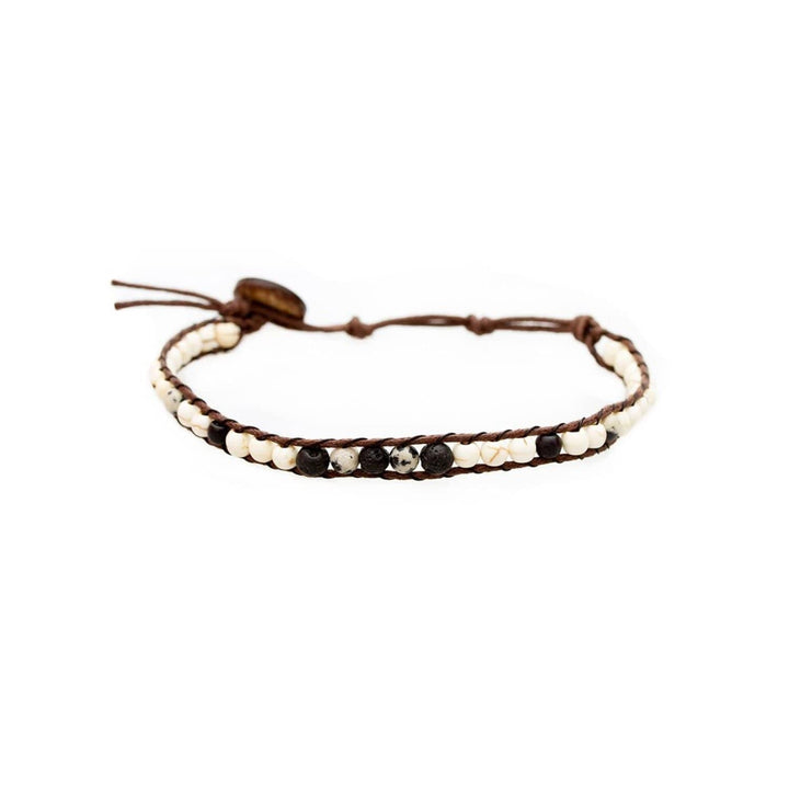 Close-up of a beaded wrap bracelet with dalmation jasper, howlite, and lava stone beads in shades of white, black, and tan with an adjustable brown cord and coconut button closure, displayed on a white bakground.