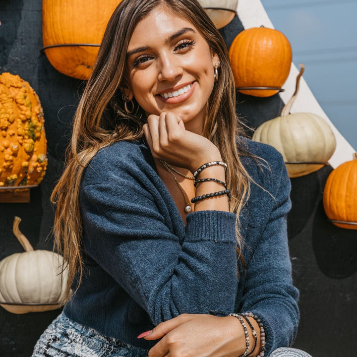 Portrait of a woman with long brown hair, a big smile with dimples, wearing a blue sweater with orange and white pumpkins in the background; she's wearing three beaded bracelets on each wrist in shades of black, white, and brwn.