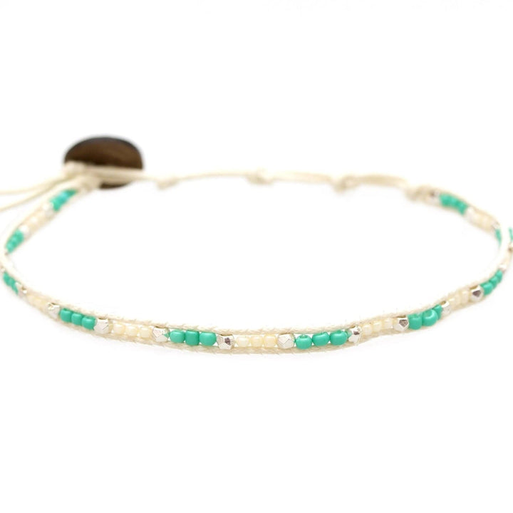 Close-up of a seed bead ankle bracelet made with turquoise, silver, creamy pearl white seed beads, with an adustable cream colored cord and a dark brown coconut shell button closure.