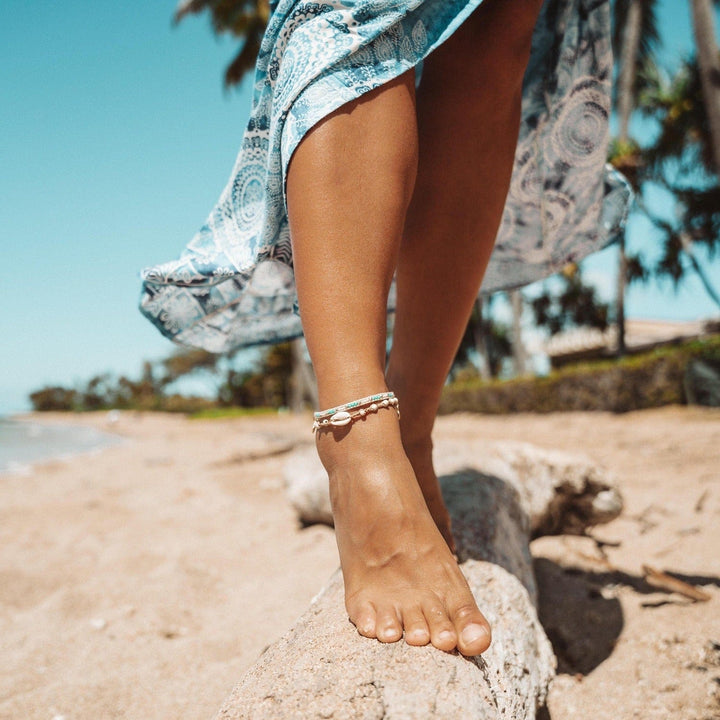 Close-up of a young woman's bare legs and feet, at the beach, standing on a tan driftwood trunk, wearing a flowy aqua blue and white printed coverup skirt, and two beaded anklets including the marine magic seed bead anklet.