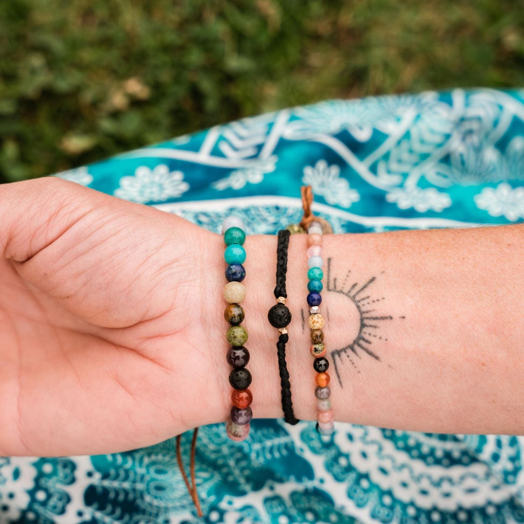 Close-up of the inside of a woman's wrist, with a black sun tattoo, wearing three bead bracelets, including the master healer bracelet, with a blurred teal print fabric and green plants background.