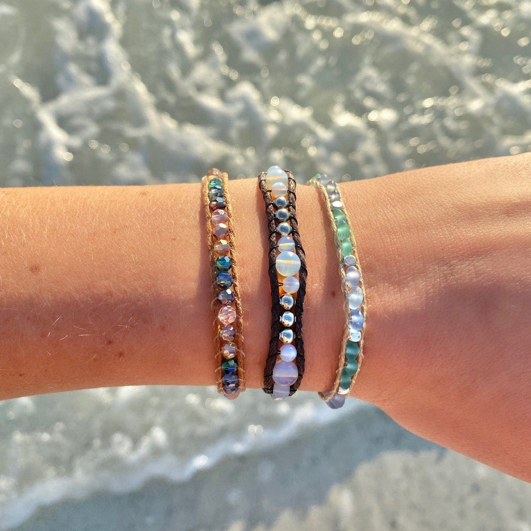 Close-up of a woman's wrist wearing three wrap style beaded bracelets in shades of pastel light blue, green, and pink, with blurred water in the background.