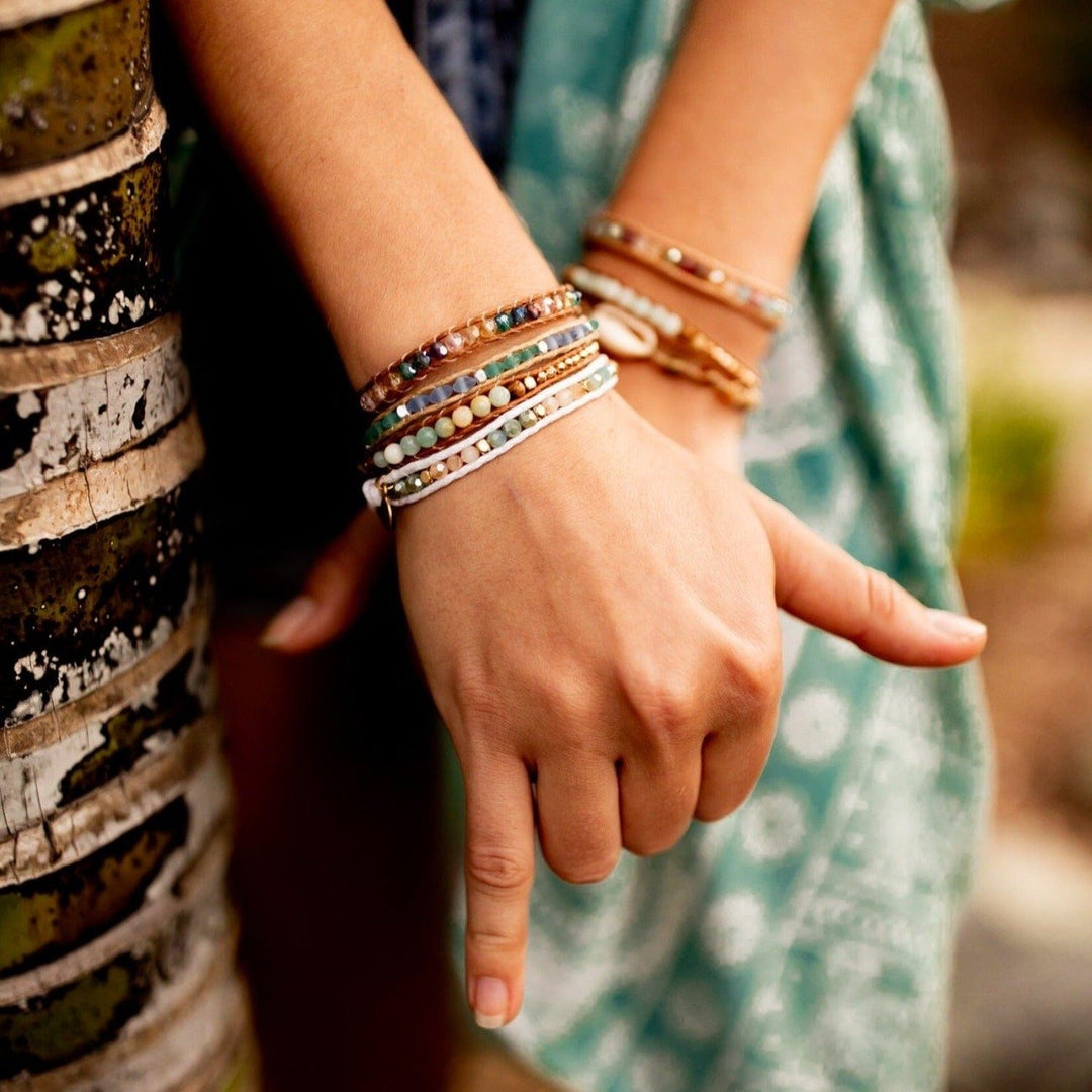 Close-up of a woman's hands crossed one over the other, nxt to a brown and tan tree trunk, wearing green printed flowy pants; she has multiple multicolored crystal bead bracelets on each wrist.