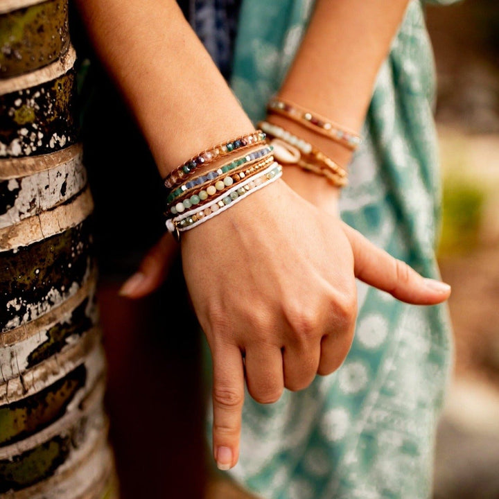 Close-up of a woman's hands crossed one over the other, nxt to a brown and tan tree trunk, wearing green printed flowy pants; she has multiple multicolored crystal bead bracelets on each wrist.