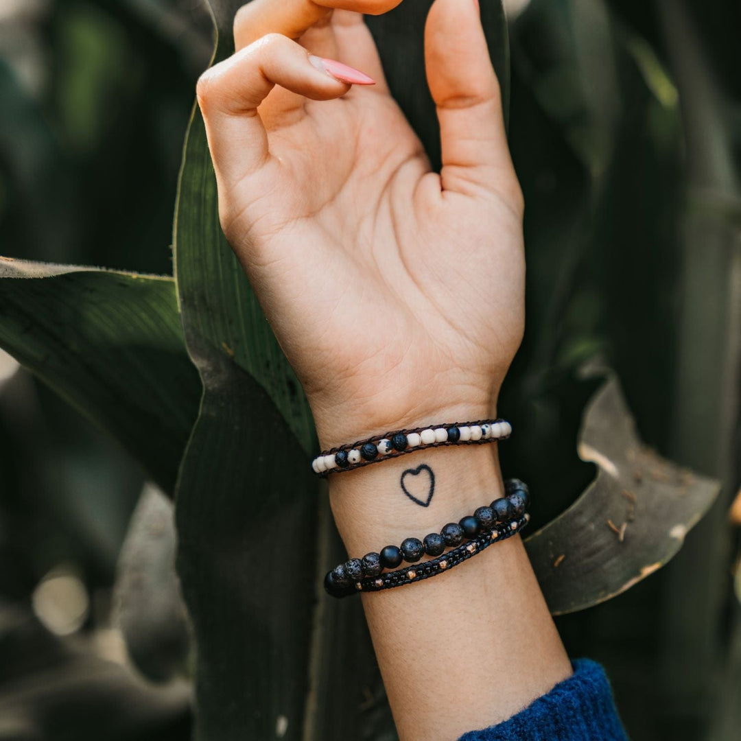 Close-up of a woman's hand and the inside of her wrist, with asmall black outline of a heart tattoo, wearing three beaded bracelets in black, white, and gold colors, with green plant leaves in the background.