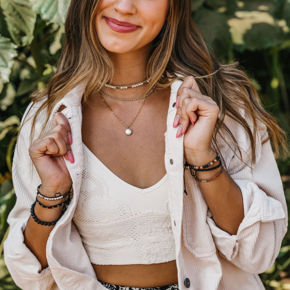 Close-up of a woman's torso, she has long brwn hair and is smiling, wearing a white top, a beaded choker necklace layered with two gold necklaces, and three beaded bracelets on each wrist in black, white, gold, brown, and green colors, with a blurred green tree in the background.