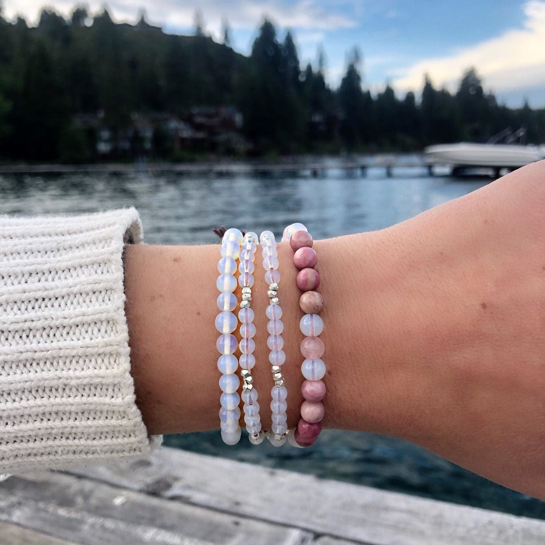 Close-up of a woman's wrist wearing four bead bracelets, including three clear blue moonstone  bracelets, and a thrid bracelet with moonstone and pink bads, with a blurred blue lake and dark green trees in the background.