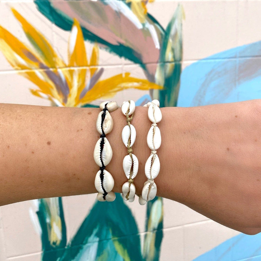 Close-up of a woman's wrist with three cowry shell bracelets, one with black cord, one with white cord, one with natural tan cord, with a wall painted with flowers in pastel colors.