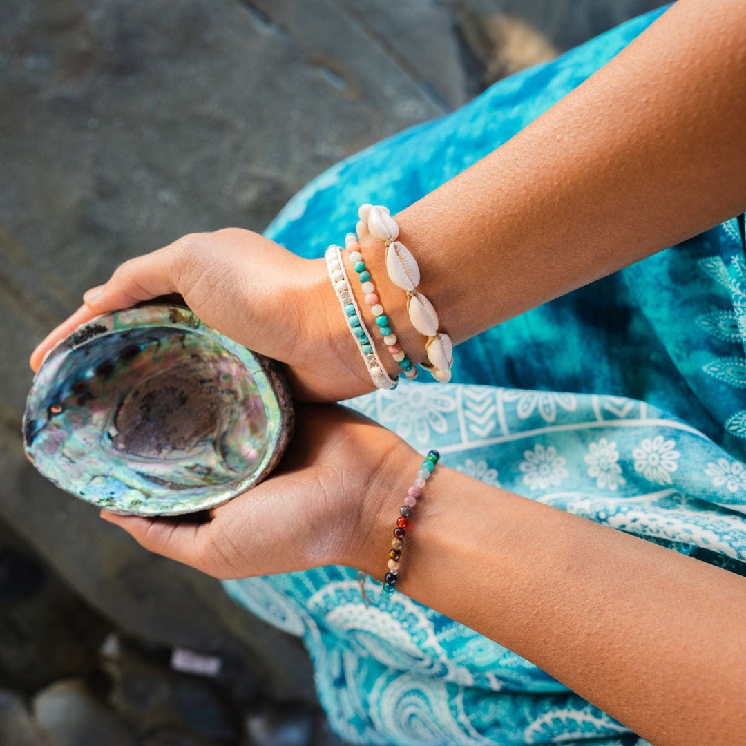 Close-up of a young woman's hands holding an abolone shell, wearing blue and white printed flowy pants, and multiple beaded bracelets on her wrists and, including a natural tan cowry shell bracelet, with gray rock background.