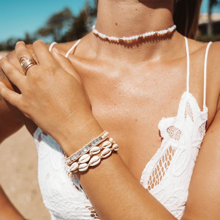 Image of a woman's chest area wearing a white sleeveless top and a white bead choker necklace, with her hand across her chest wearing a pale blue wrap bracelet, and a double wrap cowry sea shell bracelet.