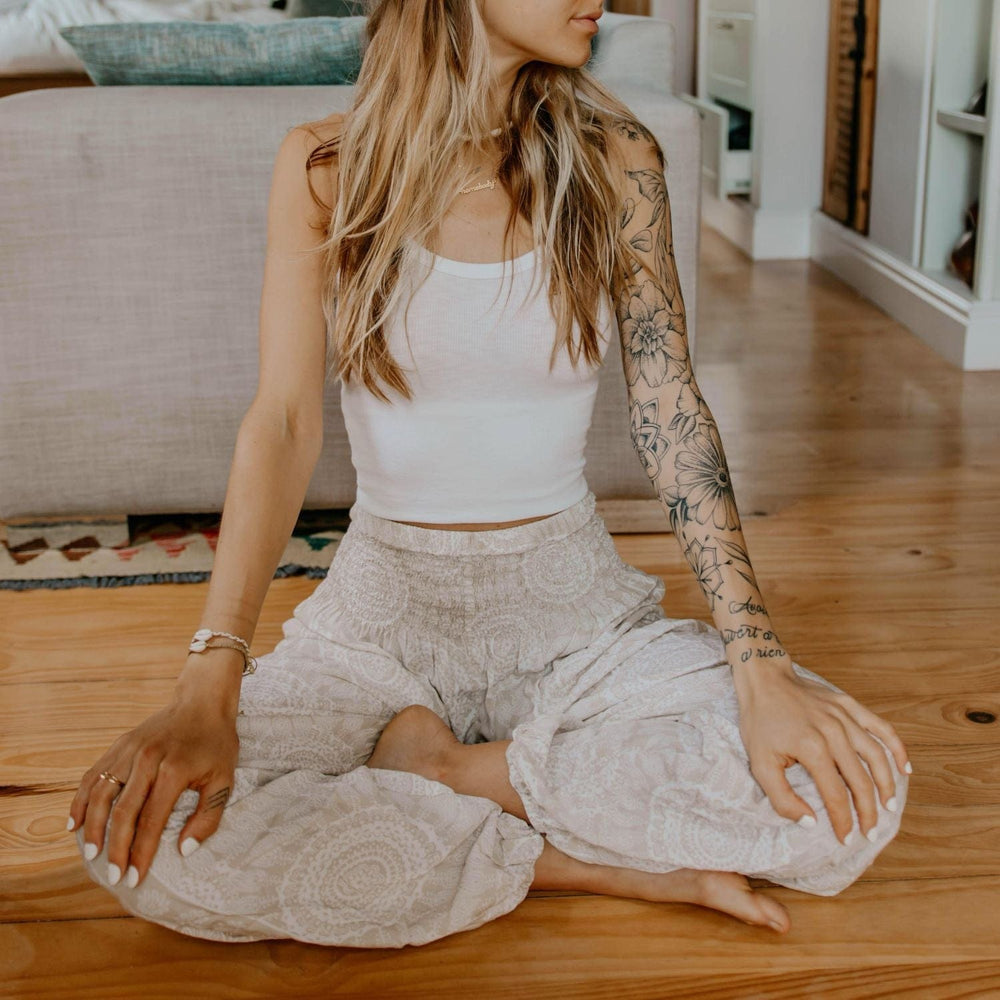 A young woman with long blond hair sitting cross legged on a golden wood floor with the back of a sofa on a rug in the background, with her hands on her knees with white fingernails, one arm has flower tattoo sleeve, wearing two tone white print flowy yoga pants and white crop tank top, wearing two braided cowry shell bracelets, including the ohana braided cowry shell bracelet.