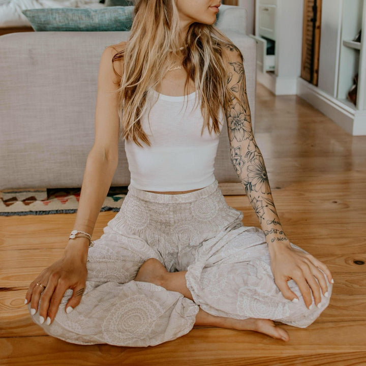 A young woman with long blond hair sitting cross legged on a golden wood floor with the back of a sofa on a rug in the background, with her hands on her knees with white fingernails, one arm has flower tattoo sleeve, wearing two tone white print flowy yoga pants and white crop tank top, wearing two braided cowry shell bracelets, including the ohana braided cowry shell bracelet.