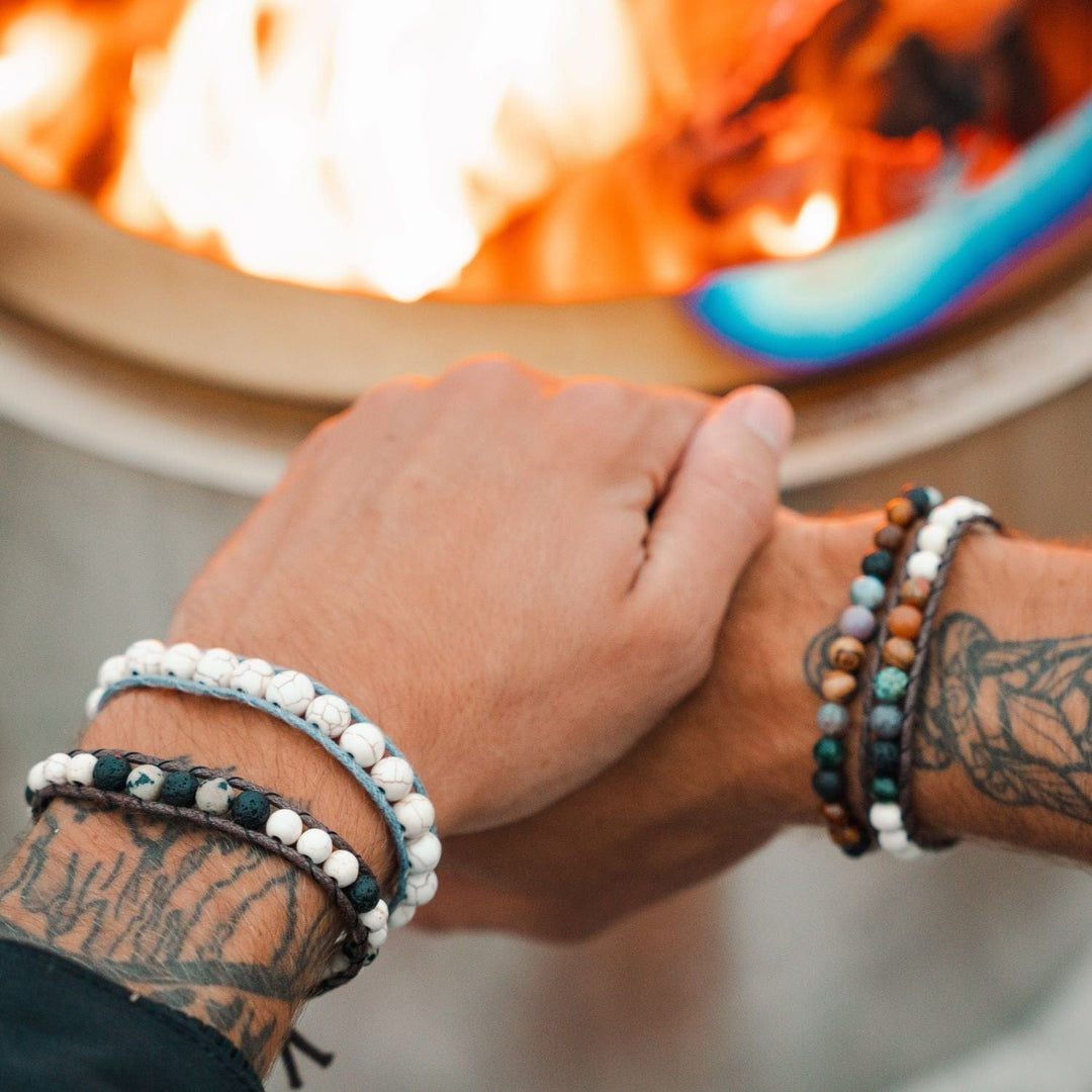 Close up of a man's hands with a tattoo two bead bead bracelets on each wrist, in black, white, brown, green, and blue colors and a blurred lit firepit in the background.