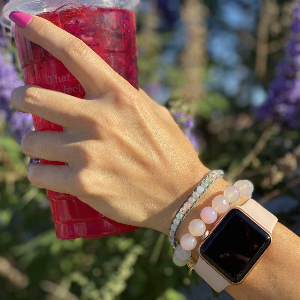 Woman's hand and wrist holding a pink drink, wearing a watch, pastel bead wrap bracelet, and pastel bead agate bracelet, with blurred green and purple plants in the background.
