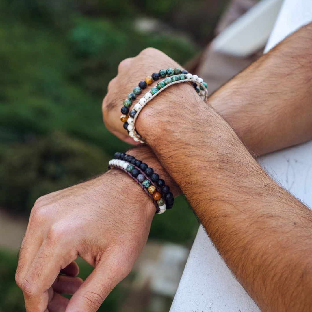 Close-up of a man's hands one crossed over the other resting on a railing, wearing two bracelets on each wrist including the peace and strength healing bracelet, with blurred green trees in background.