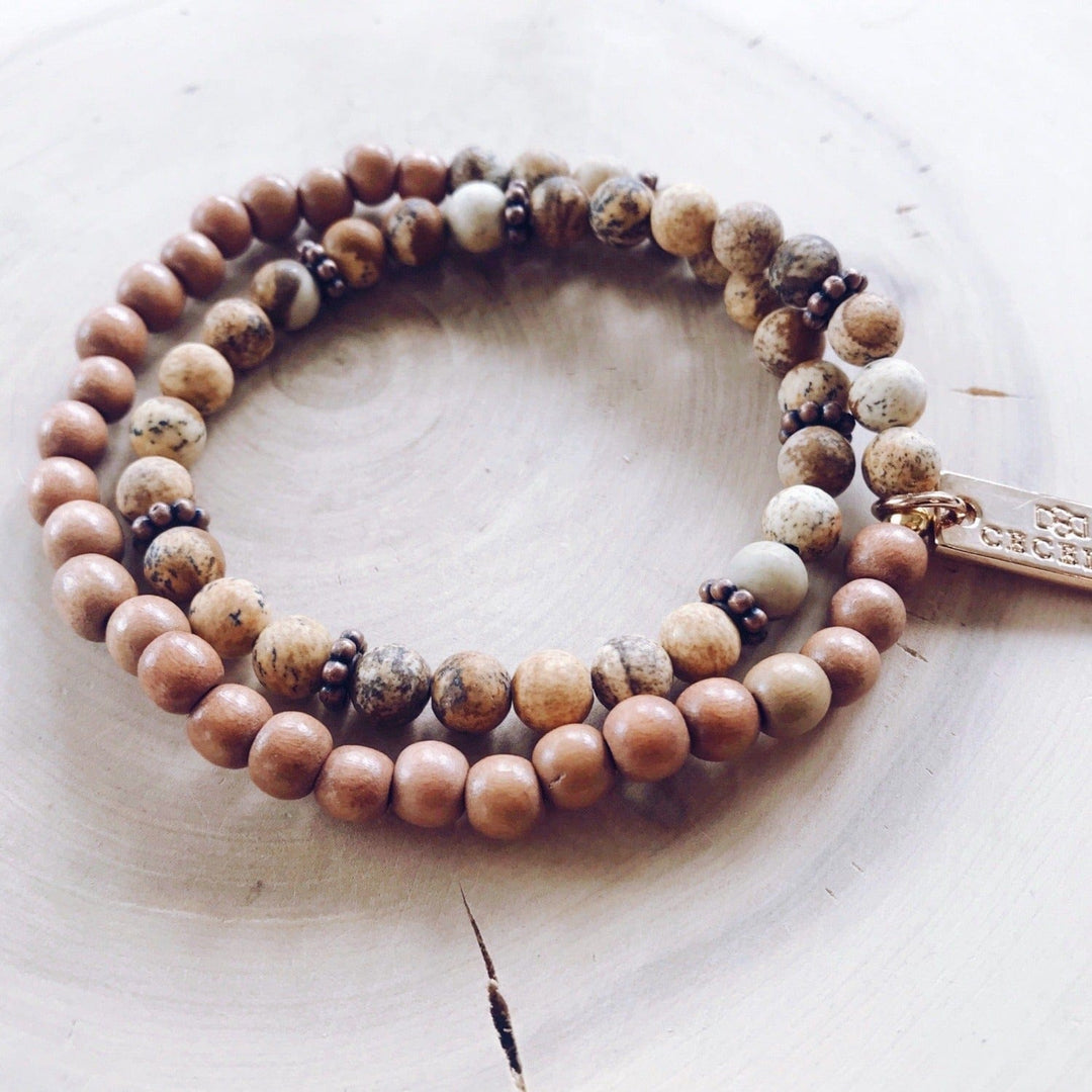 A stretch syle double-wrap bracelet made with picture jasper beads with shades of brown, and brown wood, beads with copper accent beads, and a gold-tone logo tag, displayed on a light tan background.