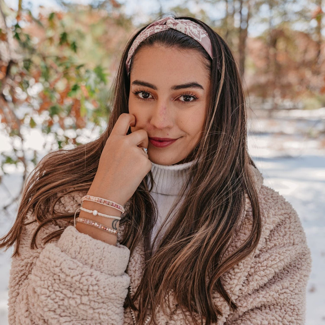 Potrait of a woman with long brown hair with a pink bandana headband, brown eys, and smiling, wearing a white sweater and a blush pink fleece winter jacket, with her hand near her face wearing three bead bracelets in shades of white and pink, with blurry white snow and brown trees in the background.