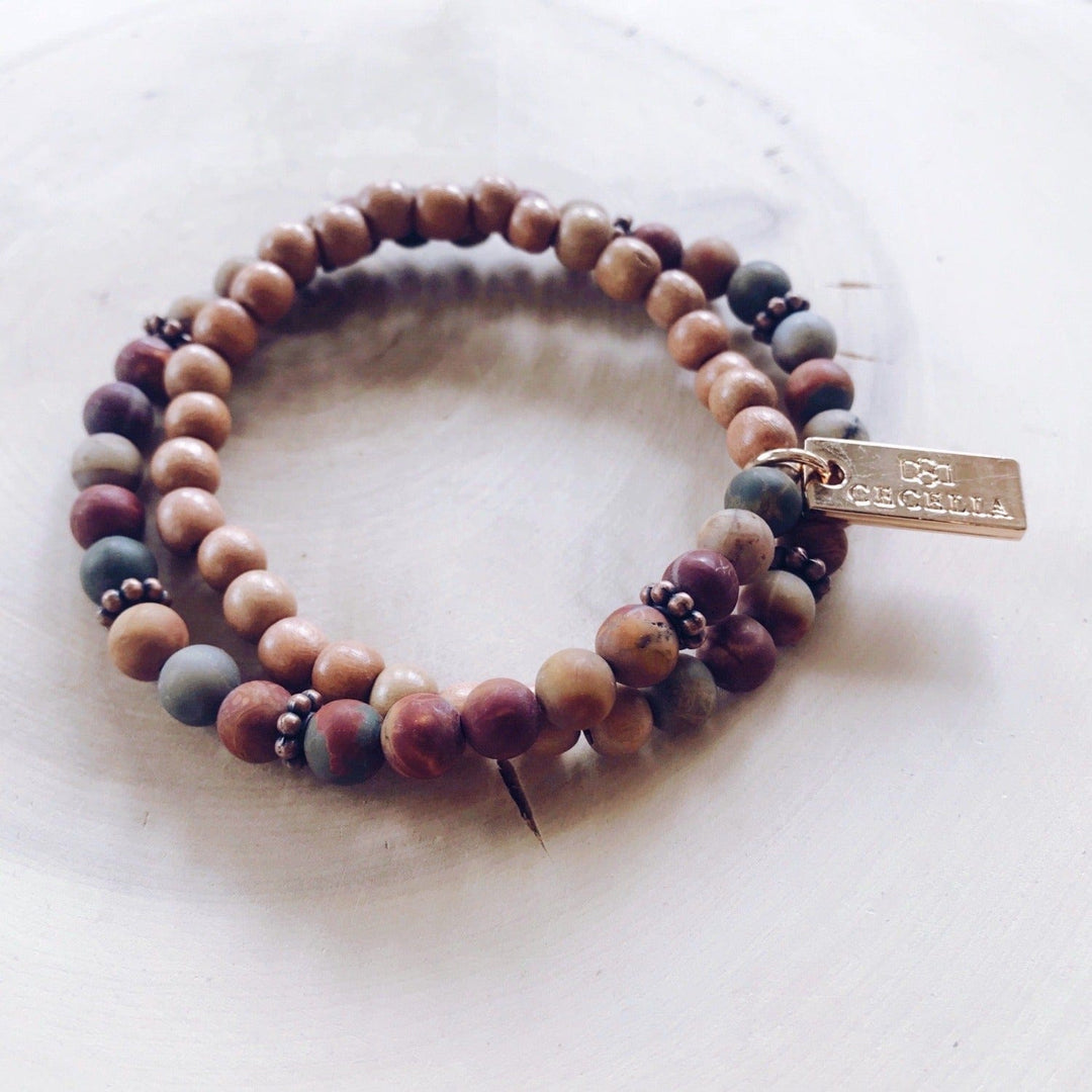 A stretch syle double-wrap bracelet made with red creek jasper beads with shades of deep red, gray, tan, and brown wood, beads with copper accent beads, and a gold-tone logo tag, displayed on a light tan background.