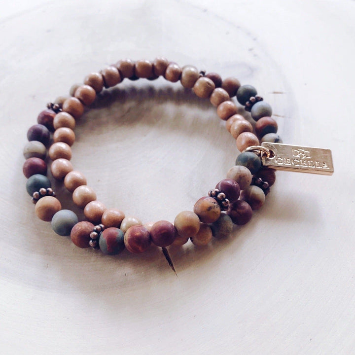 A stretch syle double-wrap bracelet made with red creek jasper beads with shades of deep red, gray, tan, and brown wood, beads with copper accent beads, and a gold-tone logo tag, displayed on a light tan background.