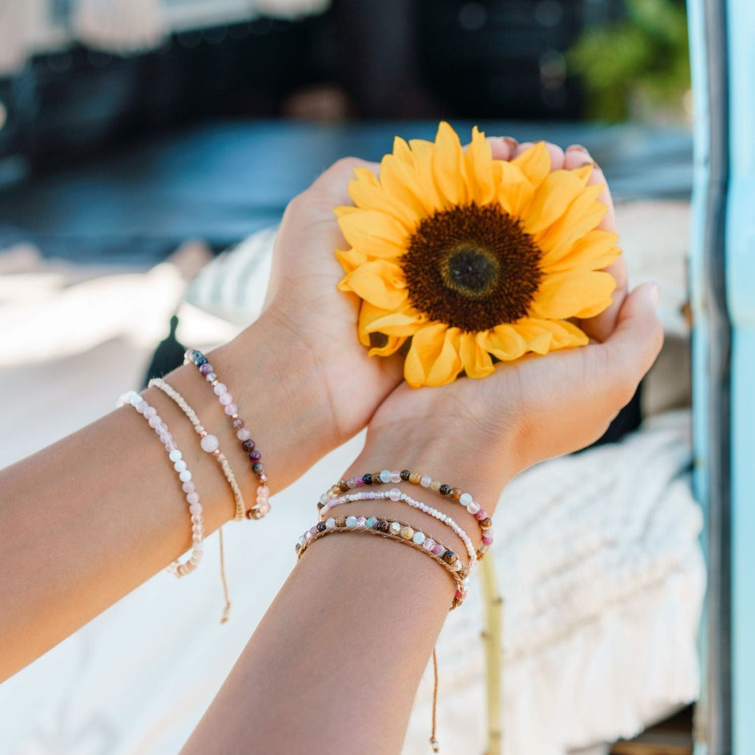 Close-up of a womans hands holding a yellow sunflower wearing three beaded bracelets on each wrist, in shades of white, brown, pink, and blue, including a rose quartz braided bracelet, with a blurred blue, white, and green background.