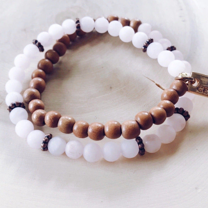 A stretch syle double-wrap bracelet made with pale pink rose quartz beads and brown wood beads, with copper accent beads, and a gold-tone logo tag, displayed on a light tan wood background.