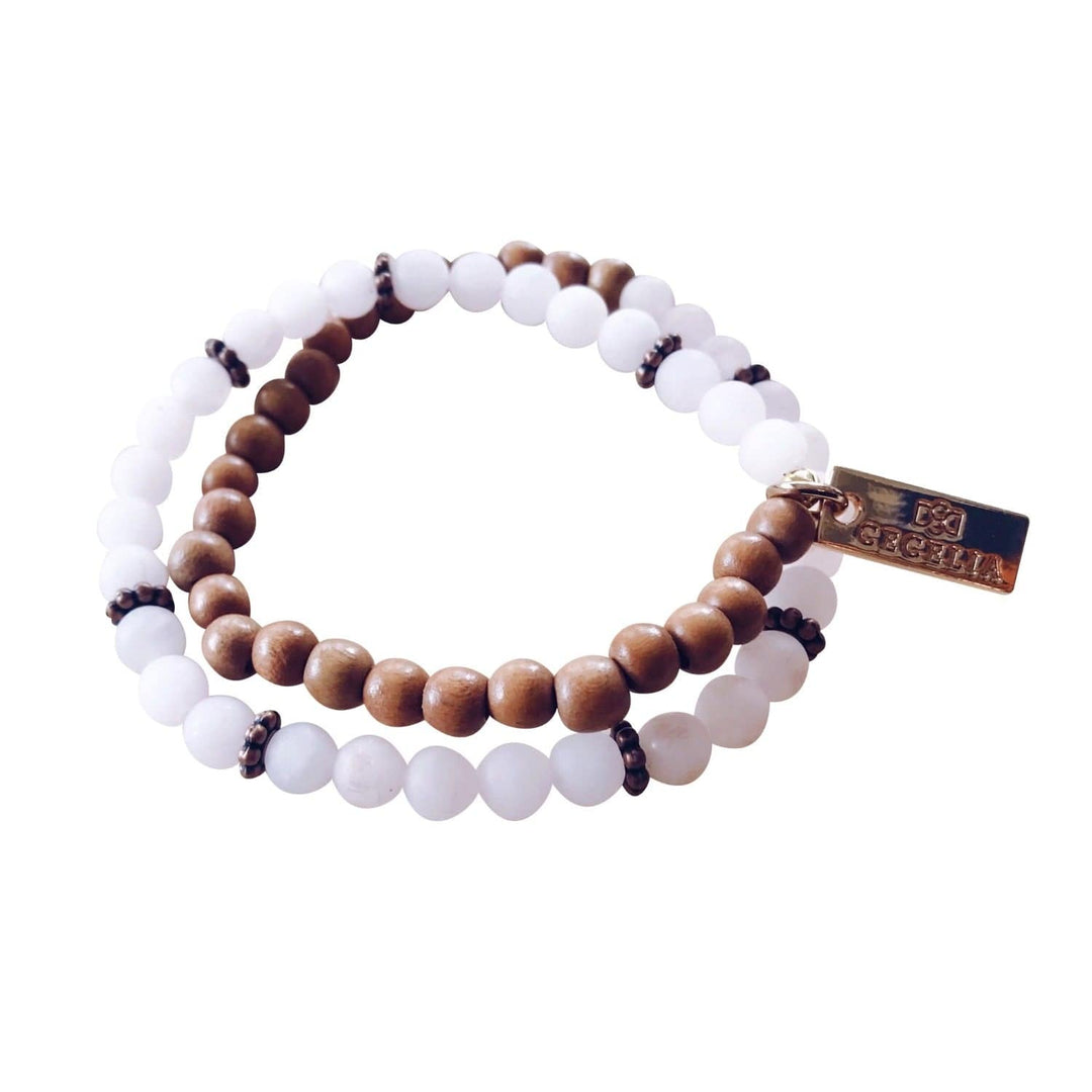 A stretch syle double-wrap bracelet made with pale pink rose quartz beads and brown wood beads, with copper accent beads, and a gold-tone logo tag, displayed on a white background.