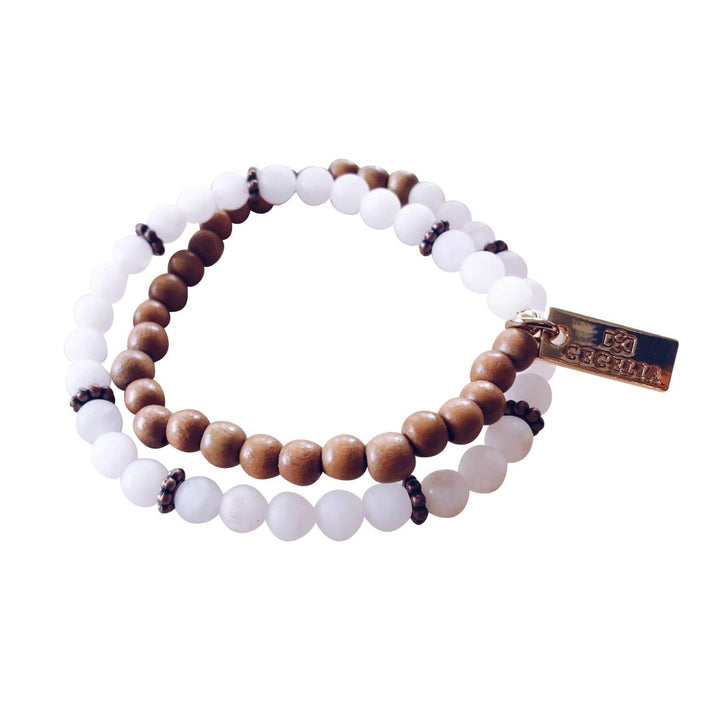 A stretch syle double-wrap bracelet made with pale pink rose quartz beads and brown wood beads, with copper accent beads, and a gold-tone logo tag, displayed on a white background.