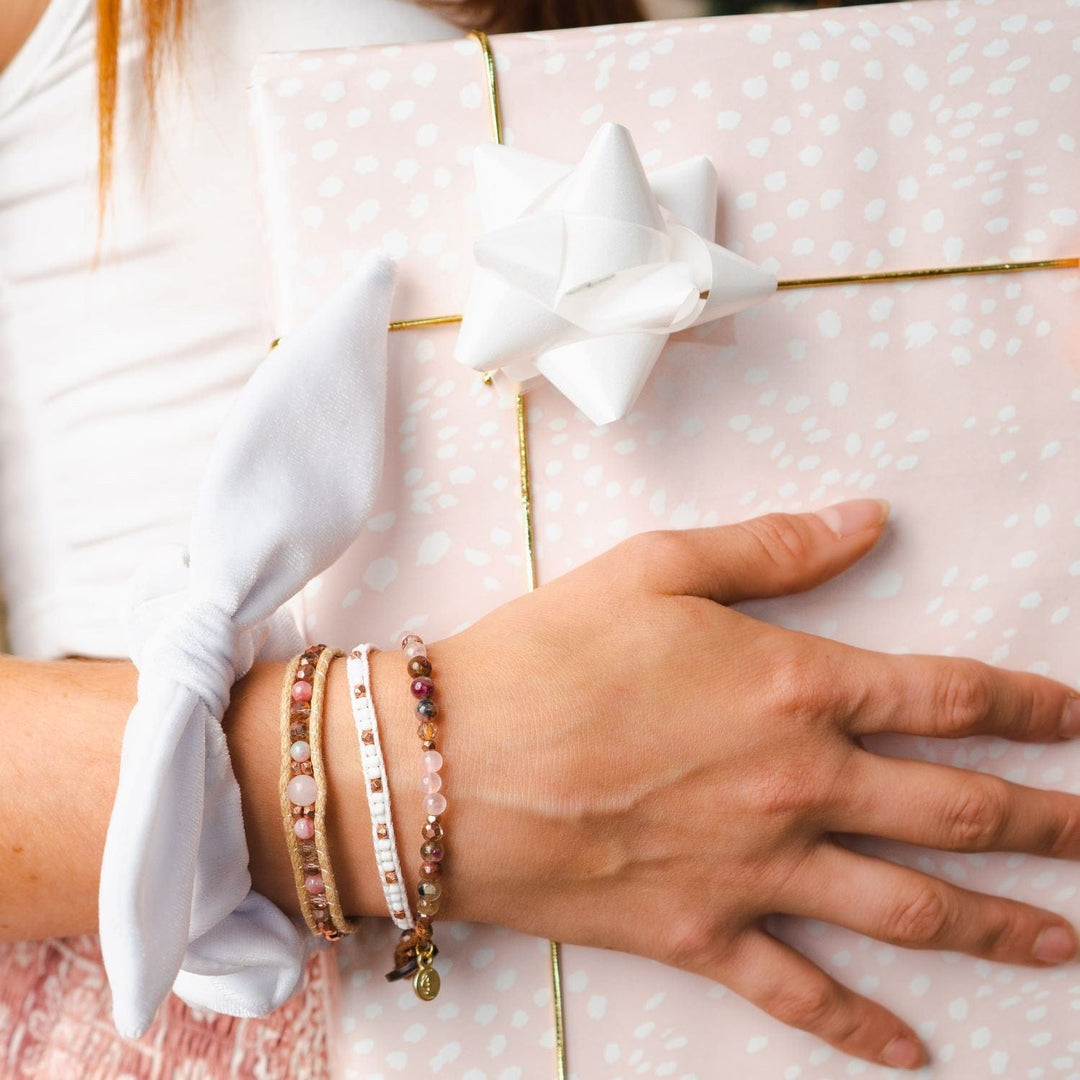 Close-up of a young woman's hand and wrist, holding a Christmas gift wrapped in pink wrapping paper with a gold cord ribbon and white bow, wearing a velvet bow scrunchie on her wrist and three beaded bracelets, in pink, white, borwn, and tan colors.