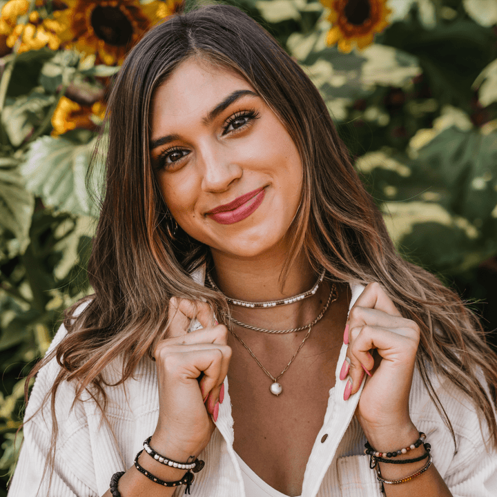Close-up image of a young woman with long brown hair smiling, wearing a white corduroy top, a beaded choker layerd with two gold necklaces, and multiple beaded wrap bracelets on each wrist, with blurred sunflowers in the background.
