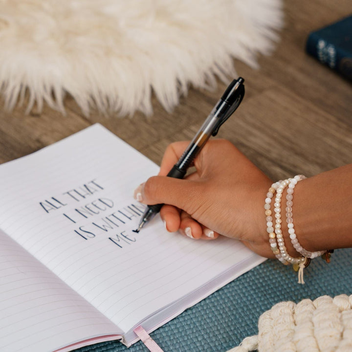 A close-up of a woman's hand holding a pen writing in a journal with text that says "All that I need is within me," wearing three bead bracelets, in white, pale yellow and pink, including a pearl bracelet, with a blurred wood floor and fur rug in the background.