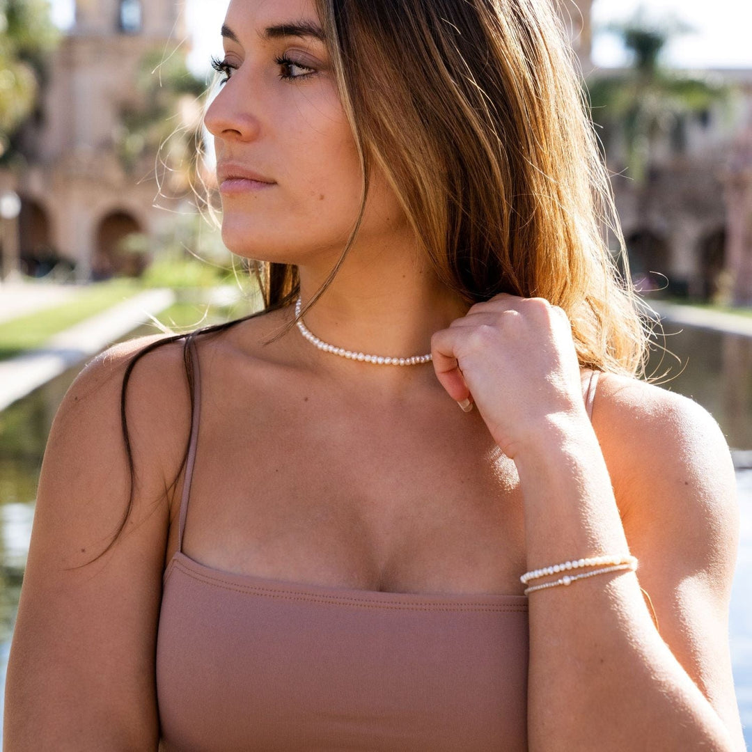 Close-up of a woman's upper body, she has long brown hair, wearing a pink tank top, a white pearl choker necklace, and two white pearl bracelets on her wrist, with blurred tan buildings and green grass in the background.
