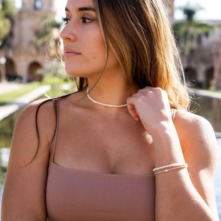 Close-up of a woman's upper body, she has long brown hair, wearing a pink tank top, a white pearl choker necklace, and two white pearl bracelets on her wrist, with blurred tan buildings and green grass in the background.