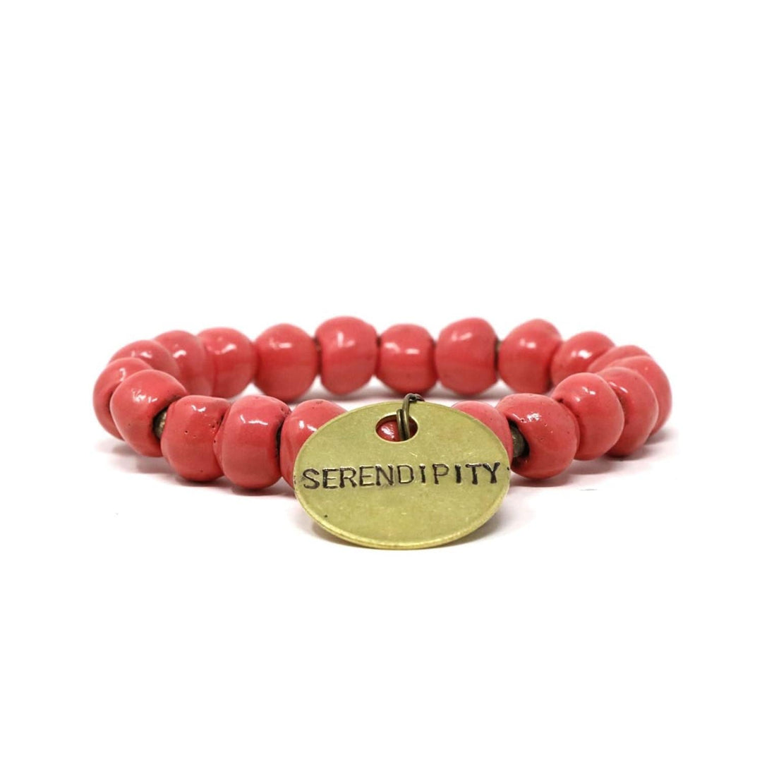 Close-up of a stretch style bracelet made with salmon red pink clay beads and gold accent beads, with a gold metal charm with the inscription 'SERENDIPITY', displayed n a white background.