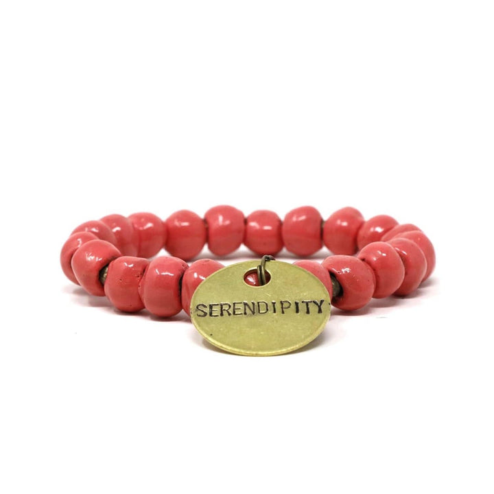Close-up of a stretch style bracelet made with salmon red pink clay beads and gold accent beads, with a gold metal charm with the inscription 'SERENDIPITY', displayed n a white background.