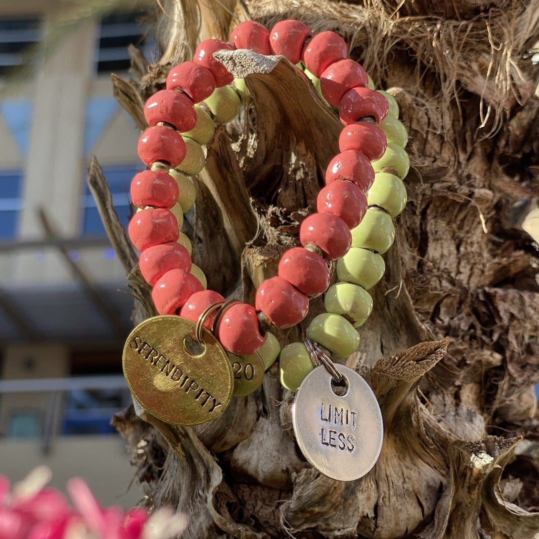 Close-up of two clay bead bracelets, one a salmon pink redcolor with gold serendipity inscribed charm, and one pastel green clay beads with silver limitless inscribed charm.