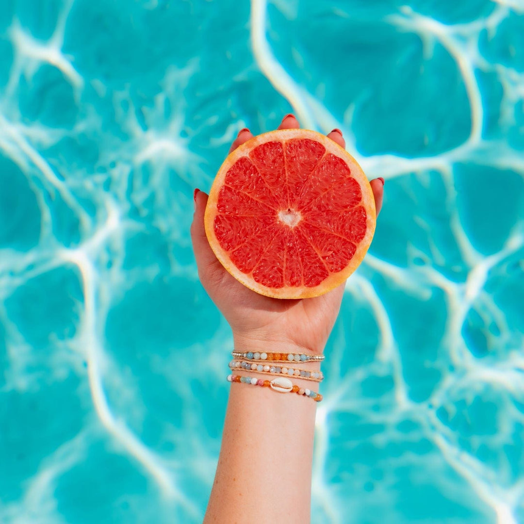 Close-up of a woman's hand holding half of a pink grapefruit with blurred blue water  in the background; she's wearing three crystal bead bracelets in shades of aqua blue, orange, blush pink, pastel green, and one has a white sea shell in the center.