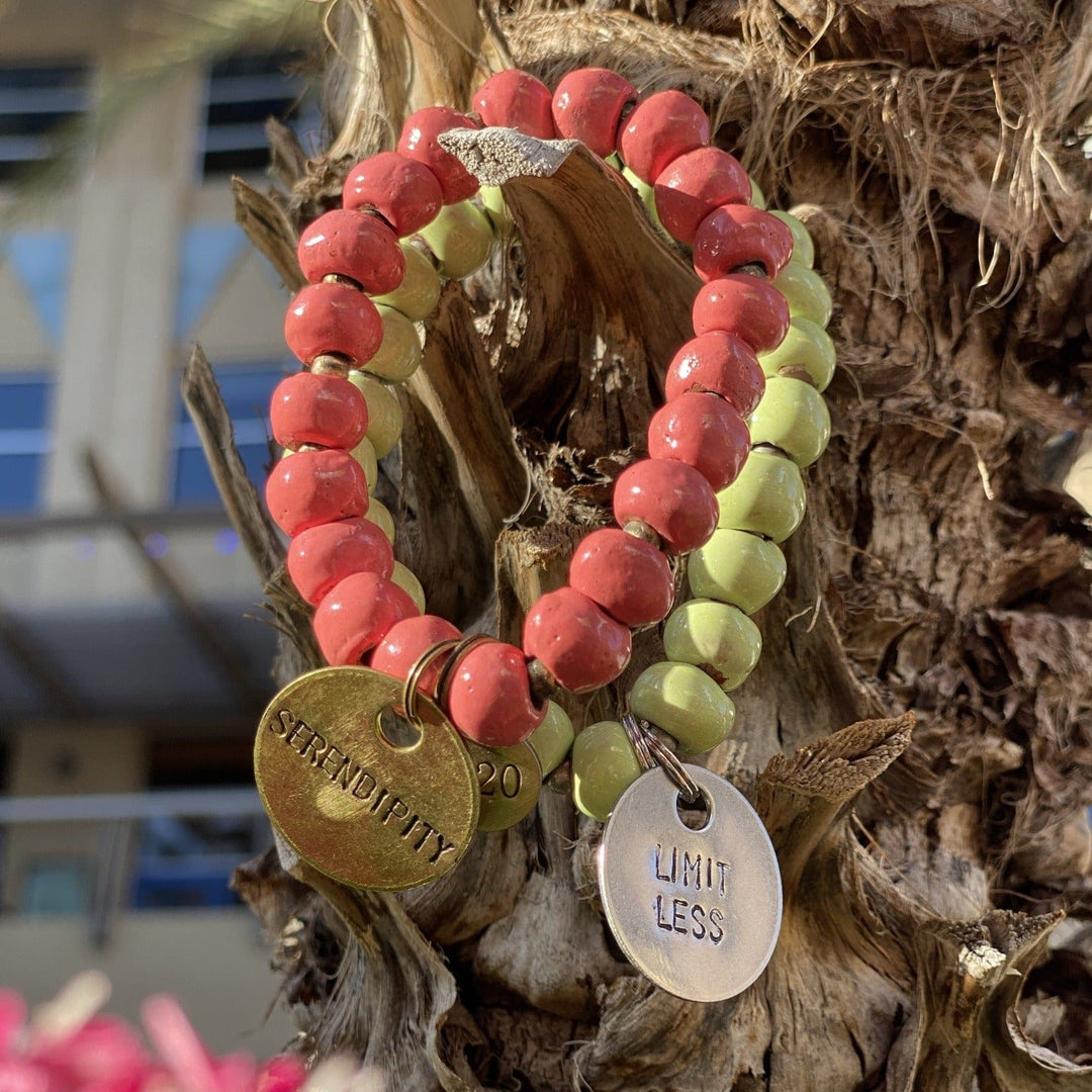 Close-up of two clay bead bracelets, one a salmon pink redcolor with gold serendipity inscribed charm, and one pastel green clay beads with silver limitless inscribed charm.