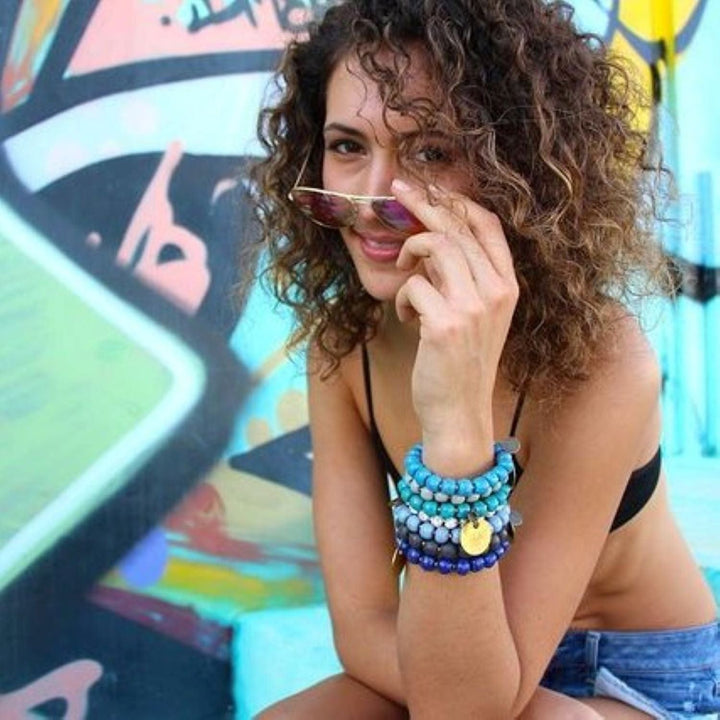 A woman with brown curly hair in a swinsuit top, denim shorts, and sunglasses; wearing seven clay bead bracelets in shades of blue and white, with gold brass inspiration charms, and a pastel color painted wall in the background.