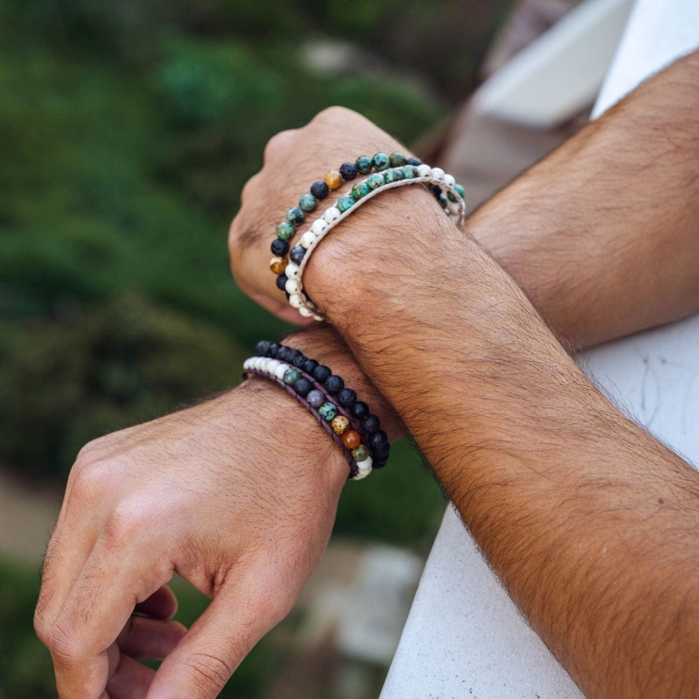 Close-up of a man's hands one crossed over the other resting on a railing, wearing two bracelets on each wrist including the strength wisdom healing bracelet, with blurred green trees in background.