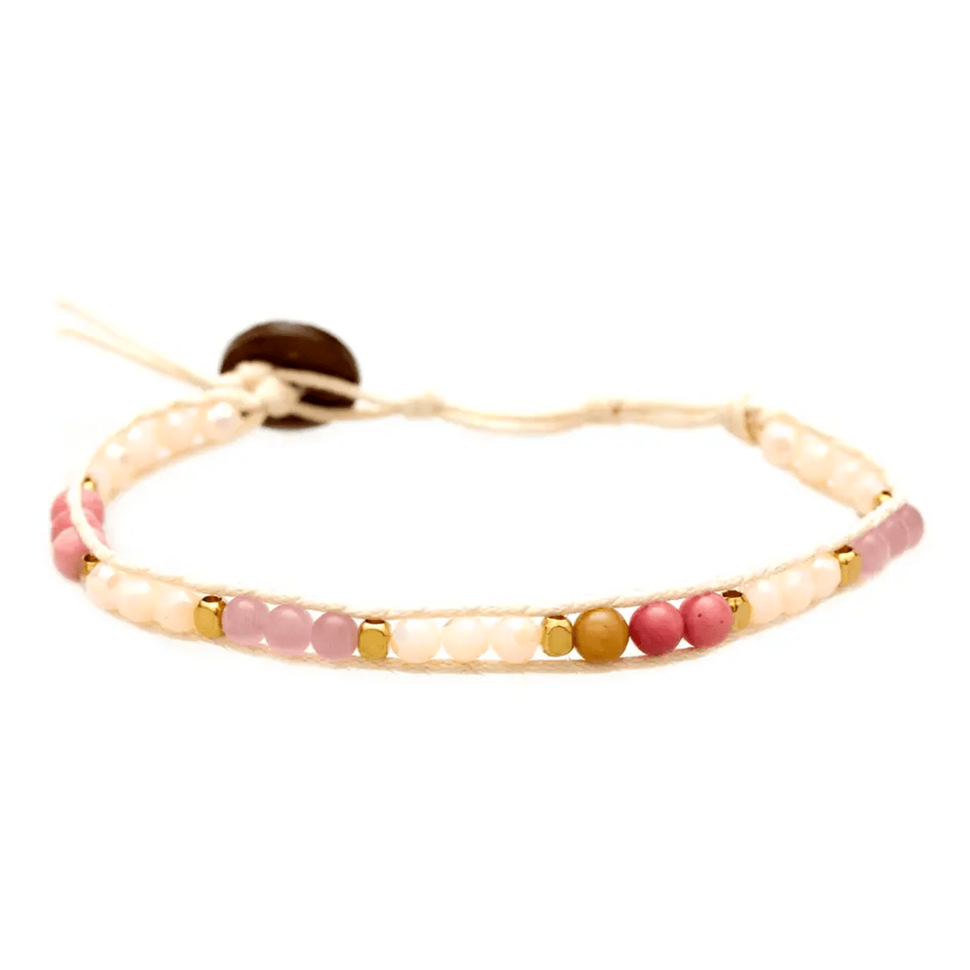 Close-up of a wrap bracelet with pink rhodolite beads, lavender beads, cream color crystals, gold accent beads, a cream white adjustable cord with a coconut button, displayed on a white background.