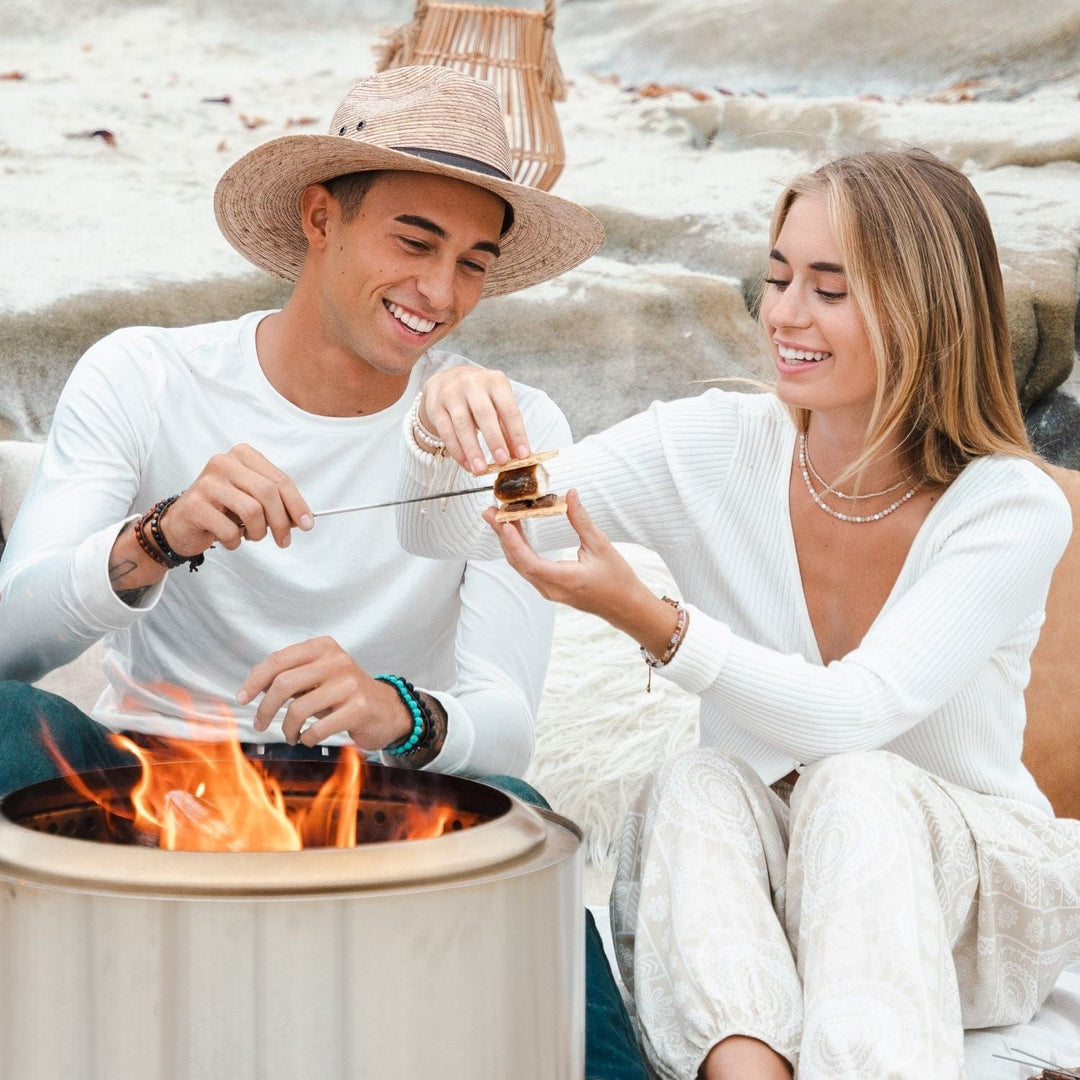 A young man and woman sitting on the beach, both wearing white clothes, making smore's over a lit firepit, both wearing beaded bracelets including the man wearing the Uluwatu cracked turquoise wrap bracelet.